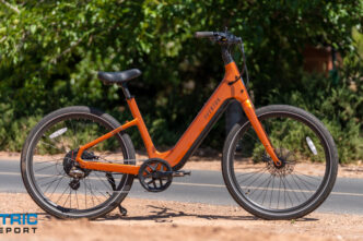 Orange electric bicycle parked on a paved path with greenery in the background and a watermark reading 'Electric Bike Report' in the lower-left corner.,