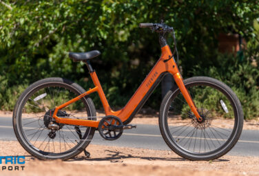 Orange electric bicycle parked on a paved path with greenery in the background and a watermark reading 'Electric Bike Report' in the lower-left corner.,