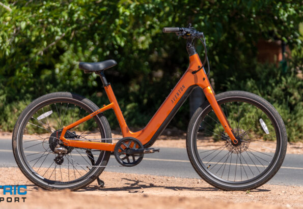Orange electric bicycle parked on a paved path with greenery in the background and a watermark reading 'Electric Bike Report' in the lower-left corner.,