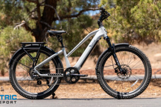 White electric bike with a step-through frame, front suspension, disc brakes, and rear rack on a paved road; Electric Bike Report logo in the bottom-left corner.