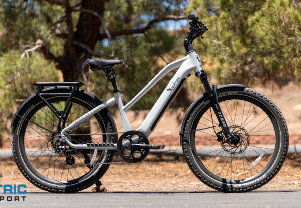 White electric bike with a step-through frame, front suspension, disc brakes, and rear rack on a paved road; Electric Bike Report logo in the bottom-left corner.