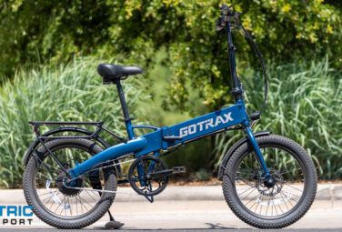 Blue GOTRAX folding electric bike parked on a paved surface with green shrubs in the background; Electric Bike Report logo in the lower left corner, image-focused product shot.