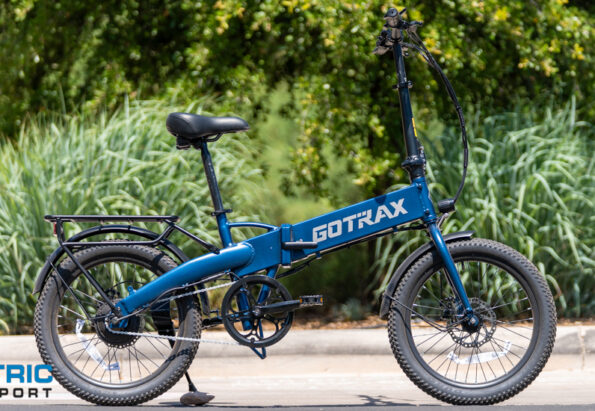Blue GOTRAX folding electric bike parked on a paved surface with green shrubs in the background; Electric Bike Report logo in the lower left corner, image-focused product shot.