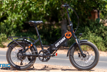 Black GOTRAX electric bike parked on a sunny roadside, side view with orange logo on the frame