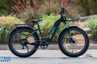 GOTRAX electric fat-tire bike parked in a paved lot with plants and a white fence in the background.