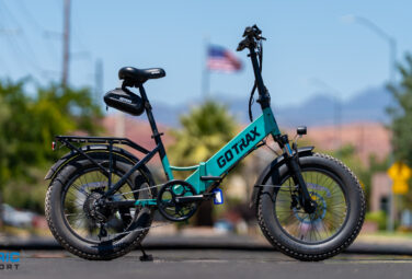 Turquoise GOTRAX electric bike with fat tires parked on a street, blurred desert town and American flag in the background.