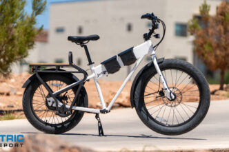 White electric bike with multiple battery packs on the frame, parked on a paved path; Electric Bike Report logo in the bottom left corner.