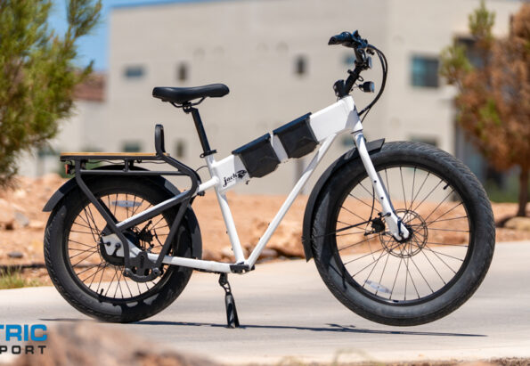 White electric bike with multiple battery packs on the frame, parked on a paved path; Electric Bike Report logo in the bottom left corner.