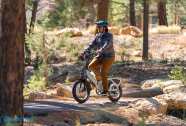 Man wearing a blue helmet and plaid shirt rides a compact electric bike through a forested trail, sunglasses on.