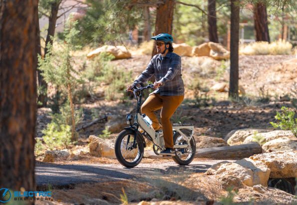 Man wearing a blue helmet and plaid shirt rides a compact electric bike through a forested trail, sunglasses on.