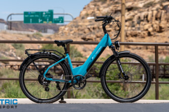 Turquoise electric bicycle parked on a bridge, desert rocks in the background, with the Electric Bike Report logo in the bottom-left corner.
