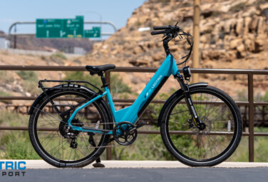 Turquoise electric bicycle parked on a bridge, desert rocks in the background, with the Electric Bike Report logo in the bottom-left corner.
