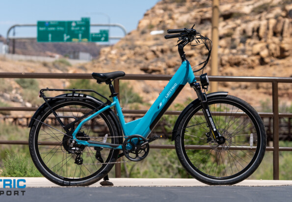 Turquoise electric bicycle parked on a bridge, desert rocks in the background, with the Electric Bike Report logo in the bottom-left corner.