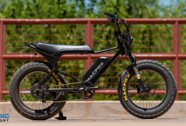 Black electric fat-tire bicycle on a bridge with a red railing; visible 'MACFOX' branding and the 'Electric Bike Report' logo in the lower-left corner.