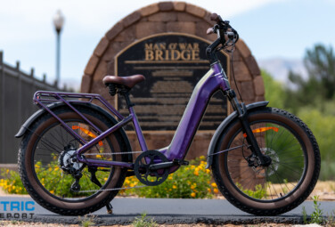 Purple electric bike with a rear rack and brown saddle parked on a path, in front of a stone arch sign that reads 'Man o' War Bridge', with an Electric Bike Report logo in the corner.