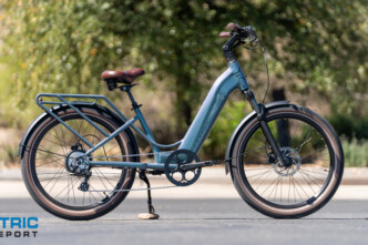 Blue step-through electric bicycle with a rear rack parked on a street, brown saddle and tires, kickstand down; Electric Bike Report logo visible bottom left.