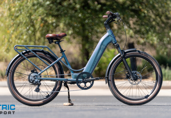 Blue step-through electric bicycle with a rear rack parked on a street, brown saddle and tires, kickstand down; Electric Bike Report logo visible bottom left.