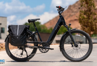 Black electric bike with a large rear pannier bag on a paved street, desert hills in the background.
