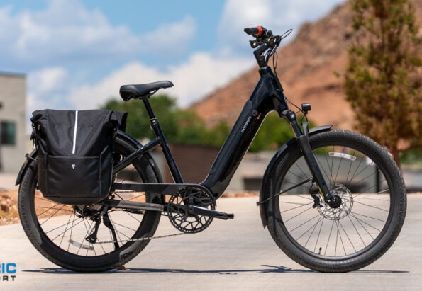 Black electric bike with a large rear pannier bag on a paved street, desert hills in the background.