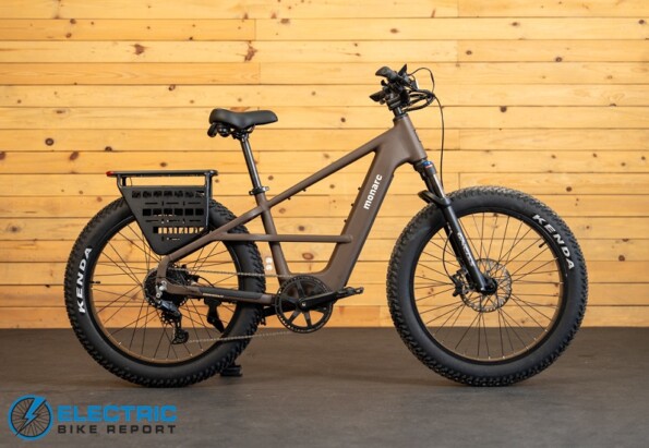 Matte brown electric bike with fat tires, front suspension, integrated battery, and a rear cargo rack against a wooden wall.