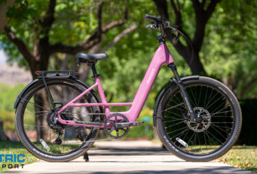 Pink electric bicycle with a black front fork parked on a sunny park sidewalk; Electric Bike Report logo in the bottom left corner.