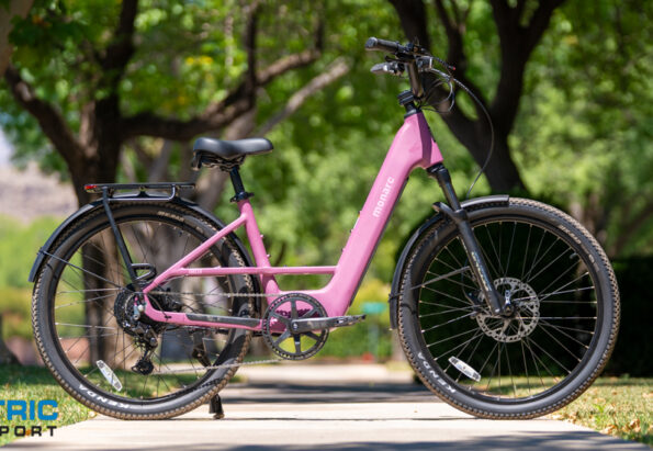 Pink electric bicycle with a black front fork parked on a sunny park sidewalk; Electric Bike Report logo in the bottom left corner.