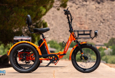 Side view of a bright orange electric bicycle with a large front rack, thick tires, and a black seat on a sunny rural path