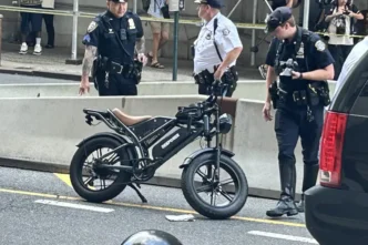 Three police officers stand near a black electric folding bike on a city street behind yellow caution tape and barriers, with bystanders in the background.