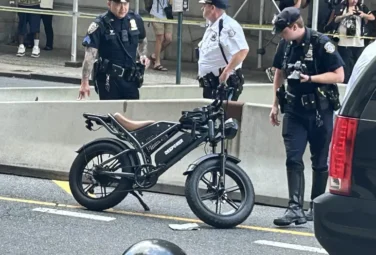 Three police officers stand near a black electric folding bike on a city street behind yellow caution tape and barriers, with bystanders in the background.