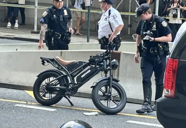 Three police officers stand near a black electric folding bike on a city street behind yellow caution tape and barriers, with bystanders in the background.