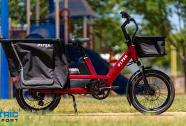 Red iFLYER electric cargo bike with large rear pannier bags parked on grass in a park; Electric Bike Report logo visible in the bottom-left corner.