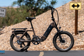 Electric bike with rear rack parked on a paved road beside a rocky slope; warning sign reads 'Do Not Enter When Flooded' and a yellow two‑way sign is in the background.