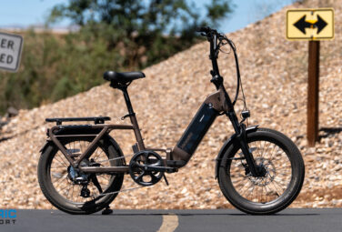 Electric bike with rear rack parked on a paved road beside a rocky slope; warning sign reads 'Do Not Enter When Flooded' and a yellow two‑way sign is in the background.
