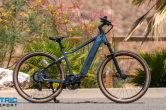 Blue mid-drive electric bike parked on a desert road; knobby tires, front suspension, and the Electric Bike Report logo in the corner.