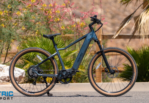 Blue mid-drive electric bike parked on a desert road; knobby tires, front suspension, and the Electric Bike Report logo in the corner.
