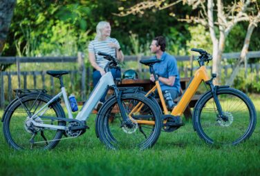 Two people chat at a wooden table in a park, with several colorful bicycles parked in front.