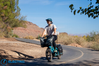 Man wearing a teal helmet rides a turquoise electric bike with a large front bag along a curving desert road, sunny day, arid scenery.
