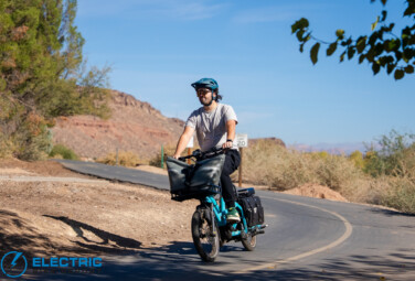 Man wearing a teal helmet rides a turquoise electric bike with a large front bag along a curving desert road, sunny day, arid scenery.