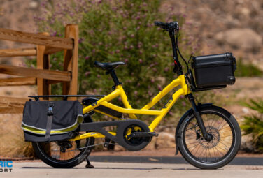 Yellow electric cargo bike with rear pannier and front storage box, parked beside a wooden fence in a desert landscape.