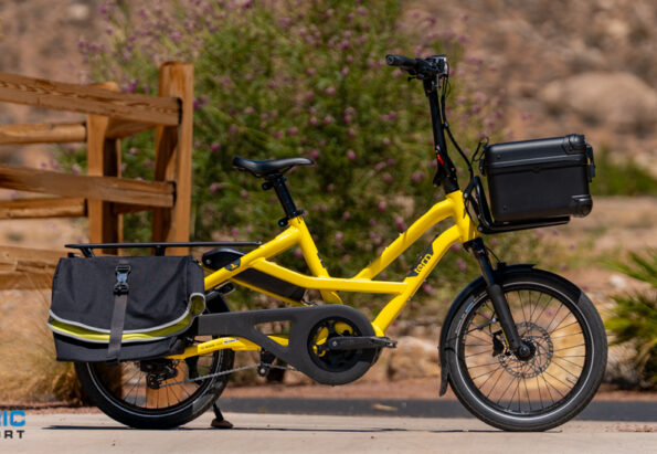 Yellow electric cargo bike with rear pannier and front storage box, parked beside a wooden fence in a desert landscape.