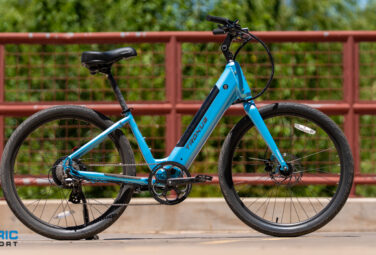 Blue Trek electric bicycle on a bridge with a red railing and green trees behind; Electric Bike Report logo bottom left.