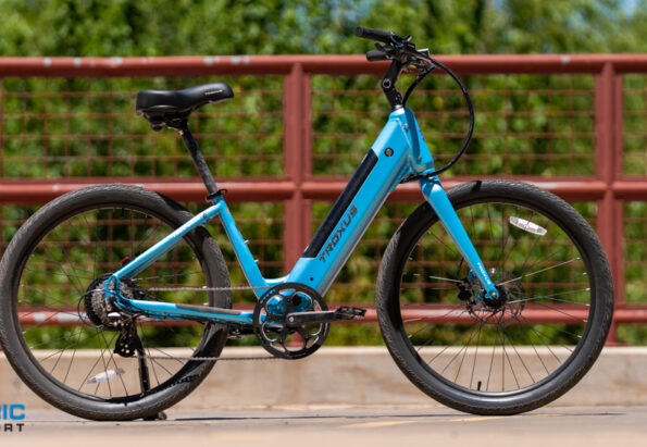 Blue Trek electric bicycle on a bridge with a red railing and green trees behind; Electric Bike Report logo bottom left.