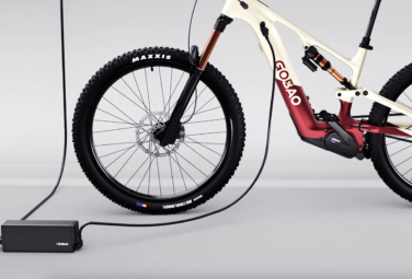 Electric mountain bike being charged, with a black charger and power cord on a light gray studio floor