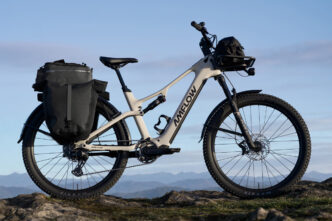 Electric mountain/gravel bike with rear panniers and a front handlebar bag parked on a rocky outcrop with distant mountains in the background, sunny sky ahead.