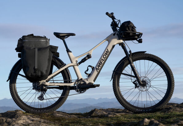 Electric mountain/gravel bike with rear panniers and a front handlebar bag parked on a rocky outcrop with distant mountains in the background, sunny sky ahead.
