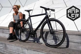 Woman in a striped tank top sits beside a black bicycle on a wooden platform, with a geometric white wall and the AMPLER logo in the background.