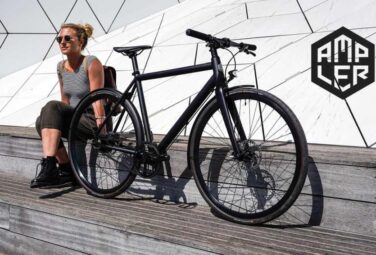 Woman in a striped tank top sits beside a black bicycle on a wooden platform, with a geometric white wall and the AMPLER logo in the background.