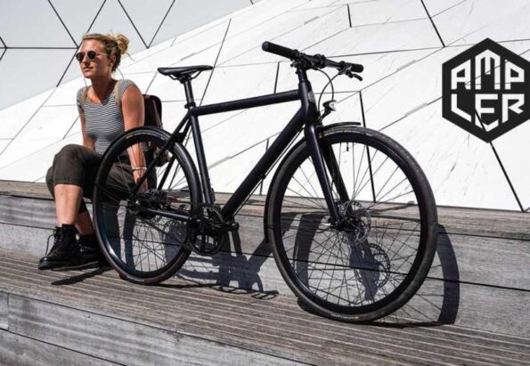 Woman in a striped tank top sits beside a black bicycle on a wooden platform, with a geometric white wall and the AMPLER logo in the background.