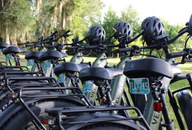Row of green rental bicycles with helmets on handlebars parked in a bike rack in a sunny park setting.