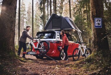Two people load bicycles onto a roof rack of a red camper van with a raised tent in a pine forest.